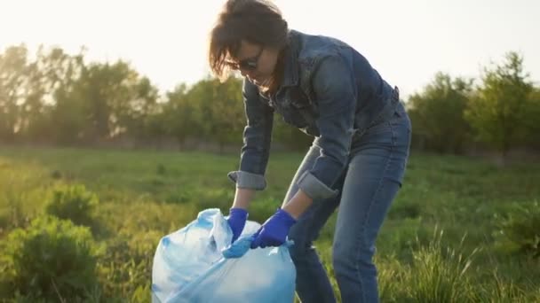 Красивая девушка на закате собирает мусор в большой пластиковый пакет. Экологическая концепция — стоковое видео