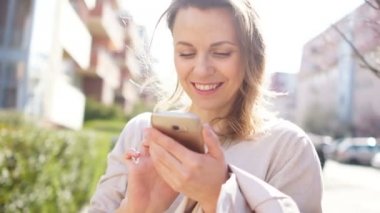 Bir şehir caddesinde akıllı telefonu olan bir kızın nazik bahar portresi, çiçekli ağaçların karşısında. Güneş parlaması, güçlü setter dalgalar saçlarını, kız gülüyor