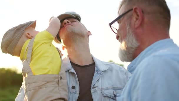 Άνδρες τρεις γενιές της ίδιας οικογένειας για μια βόλτα. Ένα χρόνο παιδί παίζει με το καπέλο του πατέρα του. Ηλιακή λάμψη — Αρχείο Βίντεο
