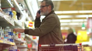 Bir süpermarkette alışveriş, telefonda konuşurken emekli. Bir adam peynir bölümünden alışveriş yapıyor. Süpermarket Arabası