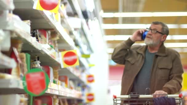 Volwassen man in de zuivel afdeling supermarkt. Gepensioneerde praat aan de telefoon en kiest kaas in de schappen van de zuivel afdeling — Stockvideo