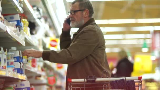 Um pensionista a fazer compras num supermercado, a falar ao telefone. Um homem faz uma compra no departamento de queijos. Carrinho de supermercado — Vídeo de Stock