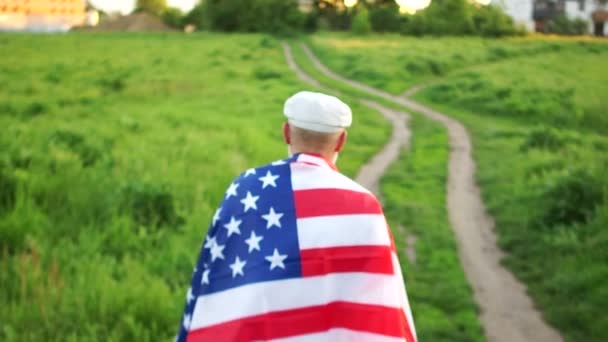 Зріла людина в шапці знаходиться на шляху до сільської місцевості. Він має Прапор США на плечах. Патріотичний день, день пам'яті загиблих американських солдатів — стокове відео