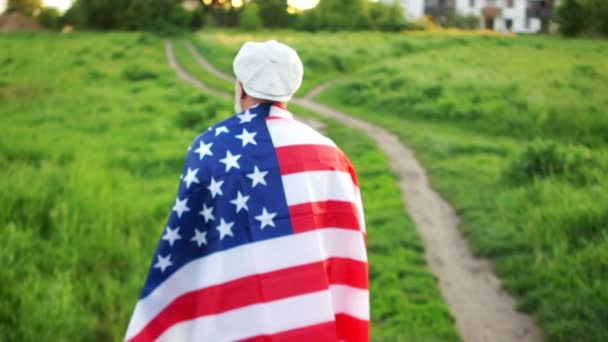 Ένας άντρας φεύγει από την πίσω πλευρά, στους ώμους της σημαίας των ΗΠΑ. Πατριωτική μέρα, η μέρα της μνήμης των νεκρών αμερικανών στρατιωτών. — Αρχείο Βίντεο