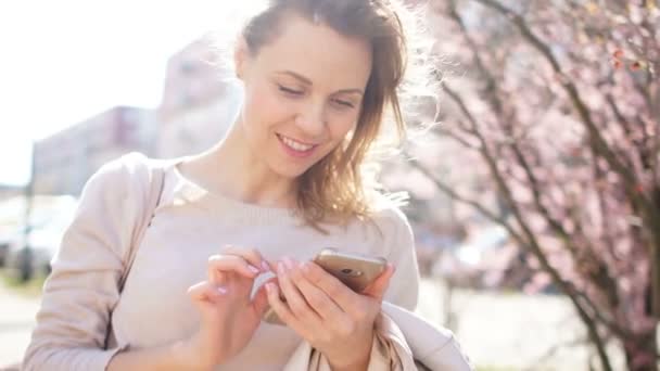 Ώριμη γυναίκα κρατώντας smartphone στα χέρια της και χαμογελώντας. Η Μανόλια ανθίζει στο δρόμο της πόλης, το ανοιξιάτικο πορτρέτο — Αρχείο Βίντεο