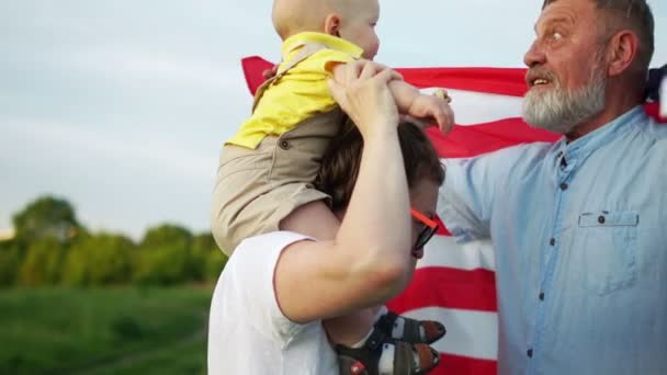 Happy American family on a picnic on July 4th. Grandfather plays with grandson. The baby sits on moms shoulders, the mans shoulders have an American symbol a flag — Stock Video