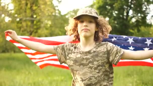 Un chico con una gran bandera americana corre por el parque. El niño está vestido con un uniforme de camuflaje y gorra. Día de la Independencia 4 de julio en los Estados Unidos — Vídeos de Stock