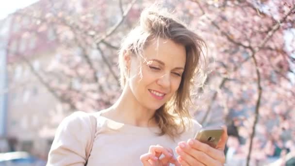 Våren porträtt av en vacker blondin med en telefon i händerna mot bakgrund av blommande Magnolia träd — Stockvideo