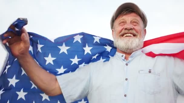 En äldre man med ett grått skägg som håller en amerikansk flagga på axlarna. Patriotiska dagen. En man i en mössa firar oss självständighets dagen den 4 juli — Stockvideo