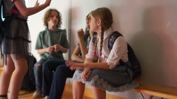 Ομάδα μαθητών με σακίδιο που κάθονται σε ξύλινο παγκάκι στο σχολικό διάδρομο. Πίσω στο σχολείο. Οι έφηβοι χαιρετά και επικοινωνούν κατά τη διάρκεια του σχολικού διαλείμματος — Αρχείο Βίντεο