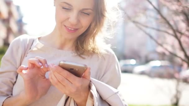 Una mujer está mirando la pantalla de su teléfono inteligente. La chica está escribiendo SMS y sonriendo mientras está de pie en medio de la calle en medio de árboles florecientes de primavera — Vídeo de stock