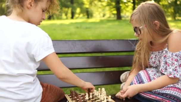 Vacaciones de verano, juegos intelectuales. Dos chicas adolescentes jugando ajedrez en un parque sentado en un banco — Vídeos de Stock