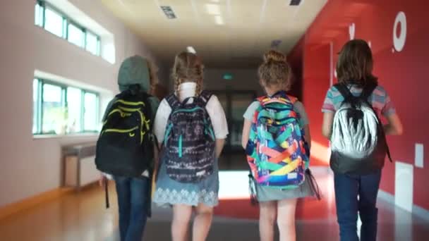 Дружественная компания подростков в школе спешит вниз по коридору к классу, вид сзади. Две девочки и два мальчика носят рюкзаки. Школьные каникулы, школьные каникулы, возвращение в школу . — стоковое видео