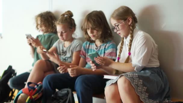 Crianças em idade escolar sentam-se em um banco na escola com seus smartphones. Vício em Internet, crianças e gadgets, de volta à escola — Vídeo de Stock