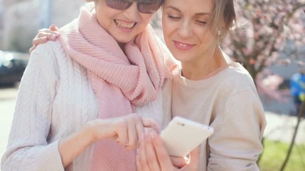Två vuxna kvinnor med en smartphone i sina händer står på gatan mot bakgrund av blommande vårträd. Diskutera foto, mors dag — Stockvideo