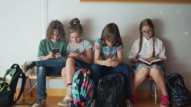 Dört öğrenci okulda bir bankta oturup akıllı telefonlarının ekranlarına bakıyor. Gözlüklü bir kız okul tatilinde kitap okuyor. Okula geri dön