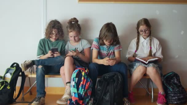 Cztery szkolne dzieci siedzą na ławce w szkole i patrzą na ekrany swoich smartfonów. Jedna dziewczyna z okularami czyta książkę podczas przerwy szkolnej. Powrót do szkoły — Wideo stockowe