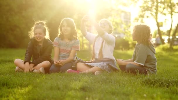 Děti ze školy sedí na trávě během školního přestávky. Krásná dívka s brýlemi a copánky dělá v paprscích zapadajícího slunce selat — Stock video