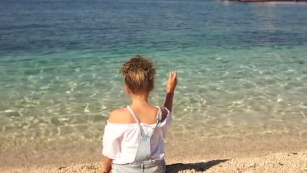 Solitaria chica rizada adolescente se sienta en la playa y arroja guijarros en el agua turquesa clara. Niños felices — Vídeos de Stock