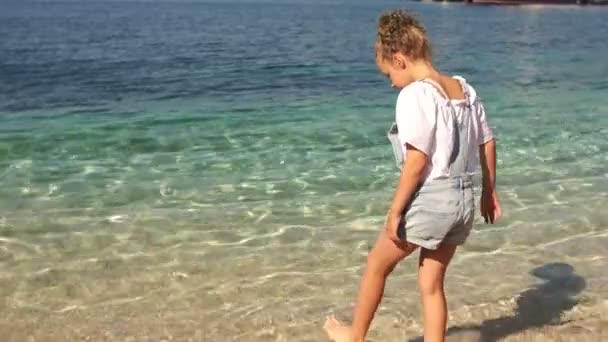 Happy kids traveling concept. Beautiful curly girl wets her feet in the clear turquoise water during the summer holidays — Stock Video