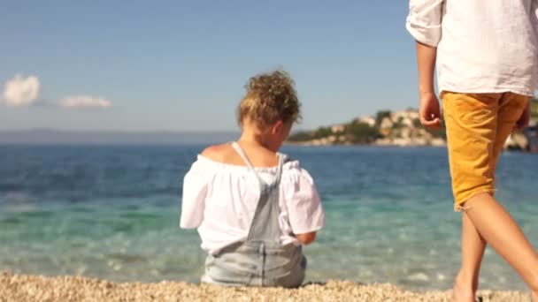 Ευτυχισμένα παιδιά κάθονται κοντά στην άκρη της θάλασσας στις ακτές της Μεσογείου. Αγόρι και κορίτσι σε φόντο διάφανου νερού από τιρκουάζ χρώμα. Σχέδιο ταξιδίου — Αρχείο Βίντεο