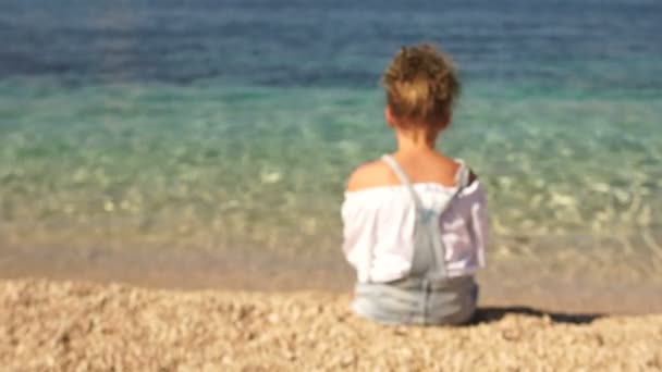 Video dinamico. La telecamera si avvicina alla ragazza seduta sulla riva del bellissimo mare limpido. Scolaretta si gira, il sole splende su di lei, bambini felici — Video Stock