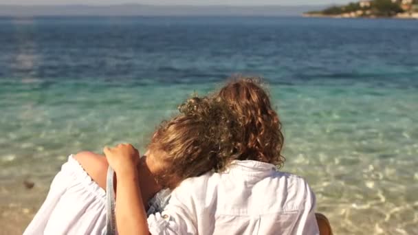 Вид сзади, кудрявый мальчик и девочка сидят обнимая на берегу самого чистого бирюзового моря, динамическая камера переводит фокус на голубые волны, концепция путешествия, счастливые дети — стоковое видео