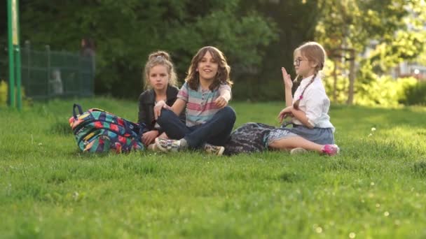 Tre compagni di classe sono seduti sull'erba durante una vacanza scolastica. Due ragazze e un ragazzo della scuola in un parco su una radura — Video Stock