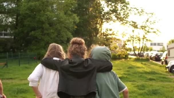 Vier klasgenoten omhelsde elkaar en liepen in de verte tegen de achtergrond van groene bomen en zonsondergang. School vriendschap, terug naar school — Stockvideo