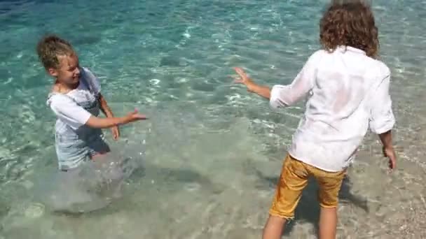 Zwei Studenten spielen im Meerwasser, ganz in Kleidern. Glückliche Kinder planschen Wasser — Stockvideo