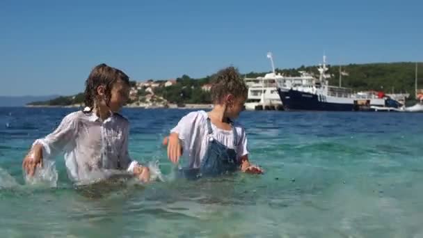 Vacaciones en Croacia. Los niños salpican en el agua de mar clara contra el fondo de una pequeña ciudad y un muelle. Hermoso paisaje y niños felices, vacaciones de verano — Vídeos de Stock