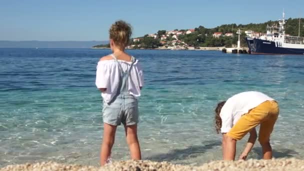 Des écoliers, un garçon et une fille, jettent des pierres à de l'eau de mer turquoise claire sur fond de ville croate et de jetée de mer avec des yachts — Video