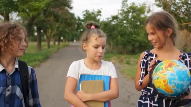 Drei Kinder lachen auf dem Weg von der Schule. trägt das Mädchen eine große Weltkugel. Wieder Schule, glückliche Kinder — Stockvideo