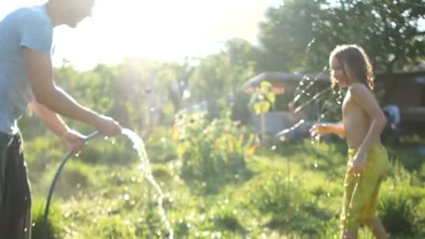 Curly-haired skolpojke åtnjuter med sin far genom att spraya varandra med vatten från en slang. Sommarvärme, semester på landet, sommarlov — Stockvideo