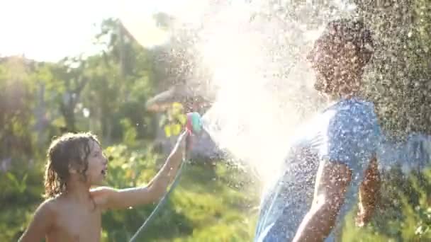 Padre e hijo se divierten en el jardín, regándose unos a otros con agua de una manguera. Spray volar alrededor, vacaciones de verano, clima caliente — Vídeos de Stock