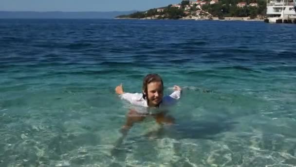 Schöne lockige Schuljunge plantscht im türkisfarbenen Meerwasser direkt in seiner Kleidung. glückliche Kindheit, Sommerurlaub — Stockvideo