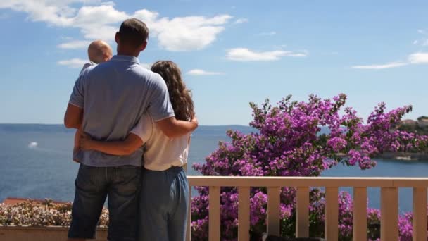 Junge Eltern mit einem einjährigen Baby auf dem Arm stehen auf einer blühenden Terrasse am Meer. Familienurlaub, Mittelmeerurlaub — Stockvideo