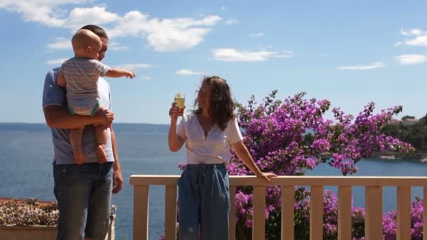 怀抱一岁婴儿的年轻父母正站在海边的一个鲜花盛开的露台上。男人带着孩子离开，母亲节，父亲节。家庭假期 — 图库视频影像