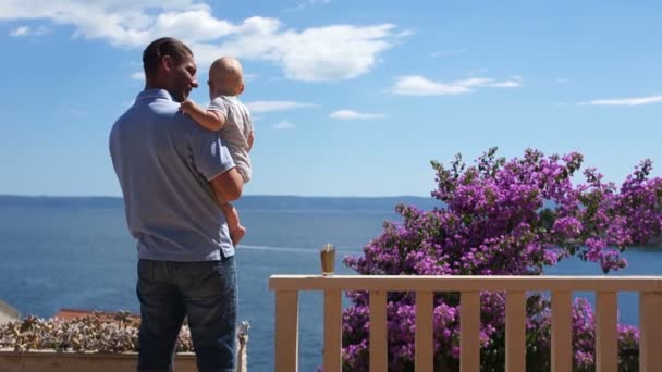 Jonge vader met zijn baby zoon in zijn armen staat op een zonnig terras aan de kust van de turquoise zee. Achteraanzicht. Fathers Day, vakantie met kinderen, gelukkig vaderschap — Stockvideo