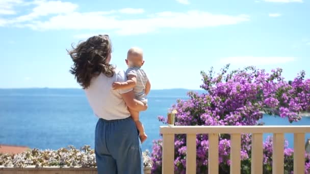 Μητέρα και παιδί στις ακτές της Μεσογείου. Μια γυναίκα με ένα παιδί στέκεται στη βεράντα και φιλάει ένα μωρό ενός έτους, οικογενειακό Σαββατοκύριακο, ημέρα της μητέρας — Αρχείο Βίντεο