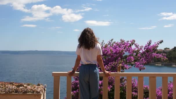 長い巻き毛を持つ若い女性が海辺のテラスに立っている。開花の茂みの近くと遠くに美しい海の景色 — ストック動画