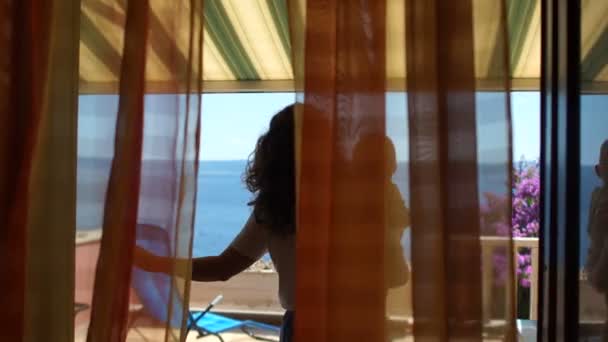 Vrouw met een baby in haar armen staat op een zonnig terras met uitzicht op zee. Resort accommodatie, familie weekend, moeder en kind — Stockvideo