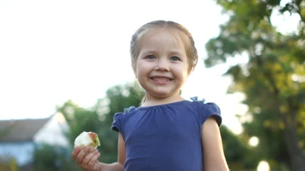 Adorable petite fille prise en gros plan à l'extérieur en été. Portrait d'une petite fille dans un village avec une pomme à la main. Vacances d'été — Video