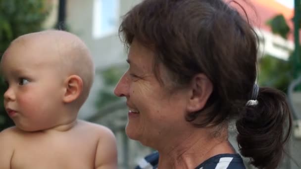 Bebé de un año en brazos de una abuela en el pueblo. Abuela al aire libre en el patio con el bebé sonriendo — Vídeos de Stock