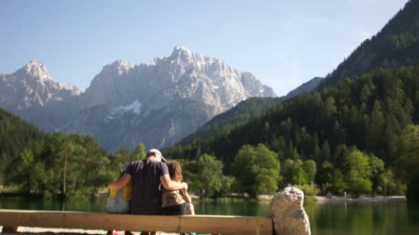 Pai e seus dois filhos adolescentes estão sentados em um banco na margem de um lago de montanha. Família admira o belo fundo da natureza. Férias de verão, férias em família felizes — Vídeo de Stock