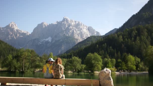 Хлопчик і дівчинка сидять на лавці на березі гірського озера на тлі гір. Літні канікули брат і сестра — стокове відео