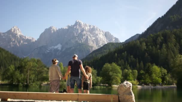 Familia de vacaciones en las montañas en el lago se divierte y juega todos juntos. Mamá y papá abrazan a su hija y su hijo, el perro corre cerca — Vídeos de Stock