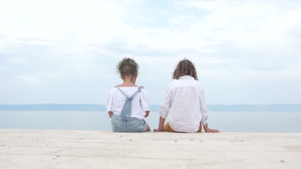 Junge und Mädchen sitzen am Ufer des klaren Meeres und blicken in die Ferne. Glückliche Kinder, reisendes Konzept — Stockvideo