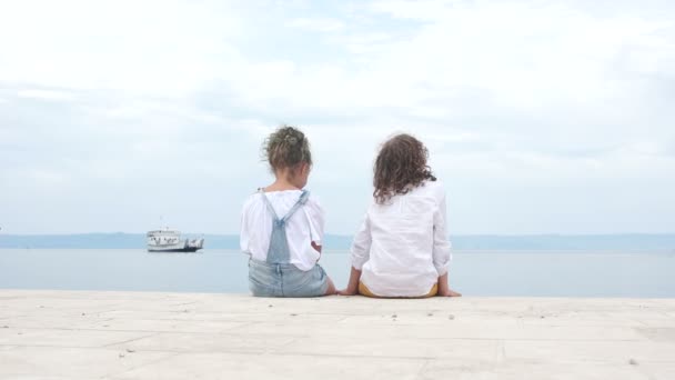 Lockenkinder, ein paar Teenager sitzen auf einem Steg am Meer und halten Händchen und schauen auf ein schwimmendes Boot. romantisches Date, erste Liebe — Stockvideo