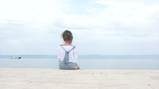 Mädchen, das allein am Ufer der Bucht sitzt und das Boot wegsieht. Ein Lockenkopf kommt herein, setzt sich neben ihn und umarmt sie. Sommerferien, glückliche Kinder — Stockvideo